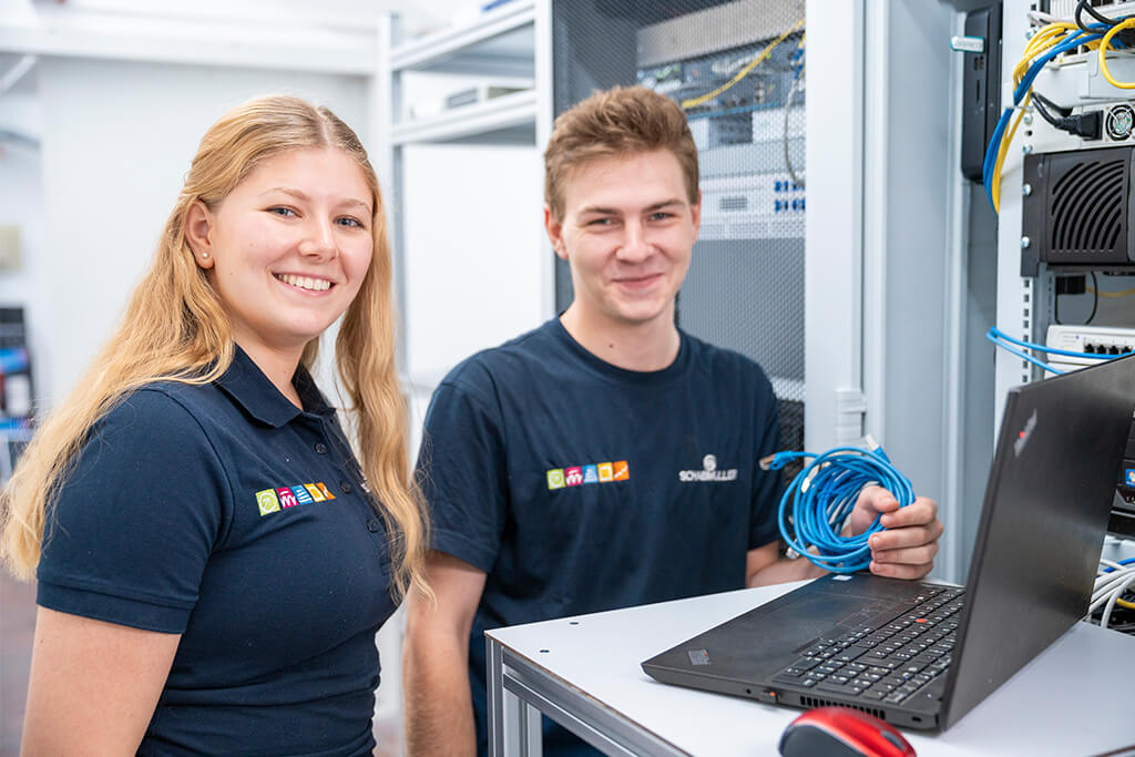 Eine Studentin und ein Student stehen bei Schabmüller am Computer und lächeln in die Kamera