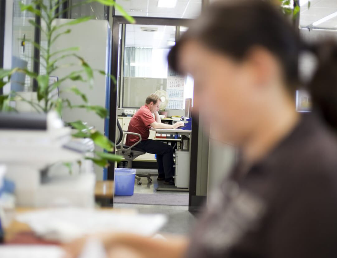 Büroaufnahme im Service von Schabmüller. Frau vorne rangezoomt verschwommen, im Hintergrund ein Mann am Schreibtisch sitzend